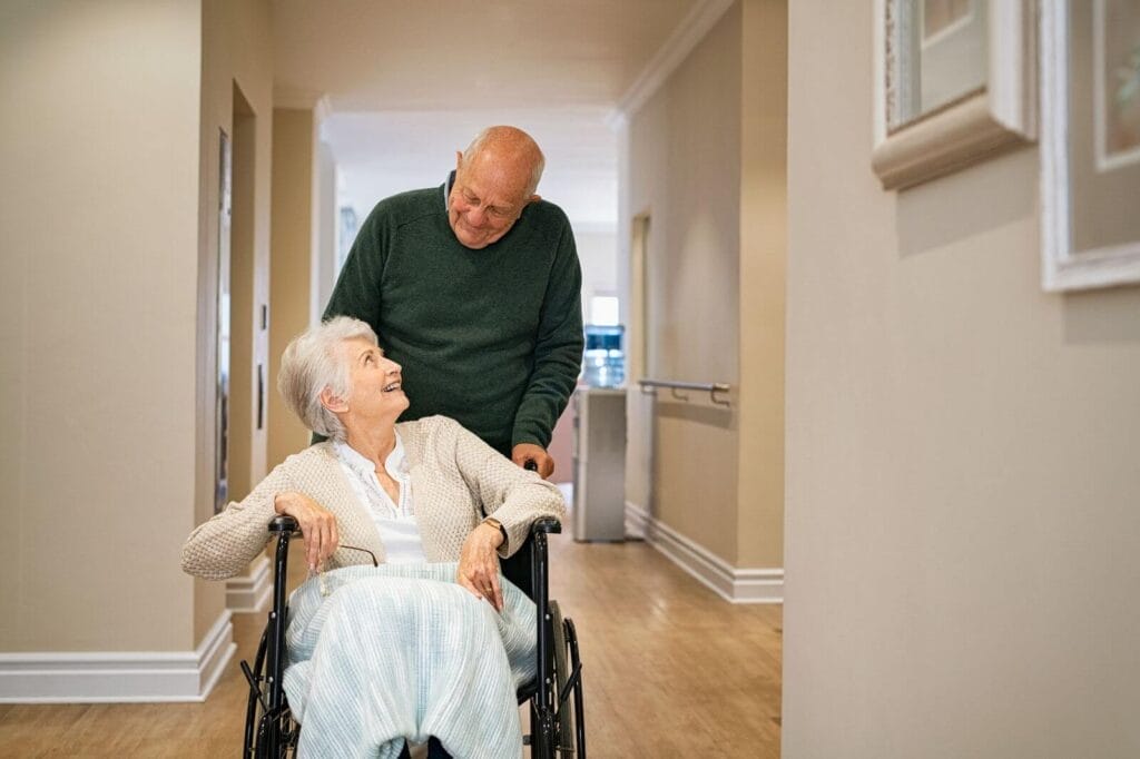 An older adult man pushes his wife in a wheelchair down a hallway. They smile at each other.