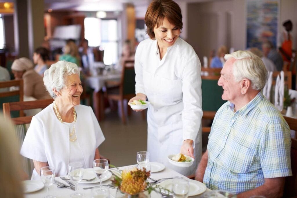 An older adult couple sits at a table in a room filled with others. A woman is placing food in front of them.