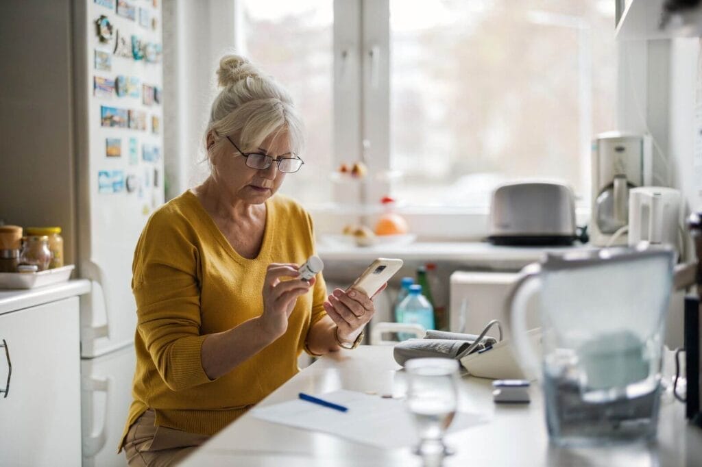 An older adult woman sits at her kitchen counter. She is looking at a medication bottle and holding her cell phone.