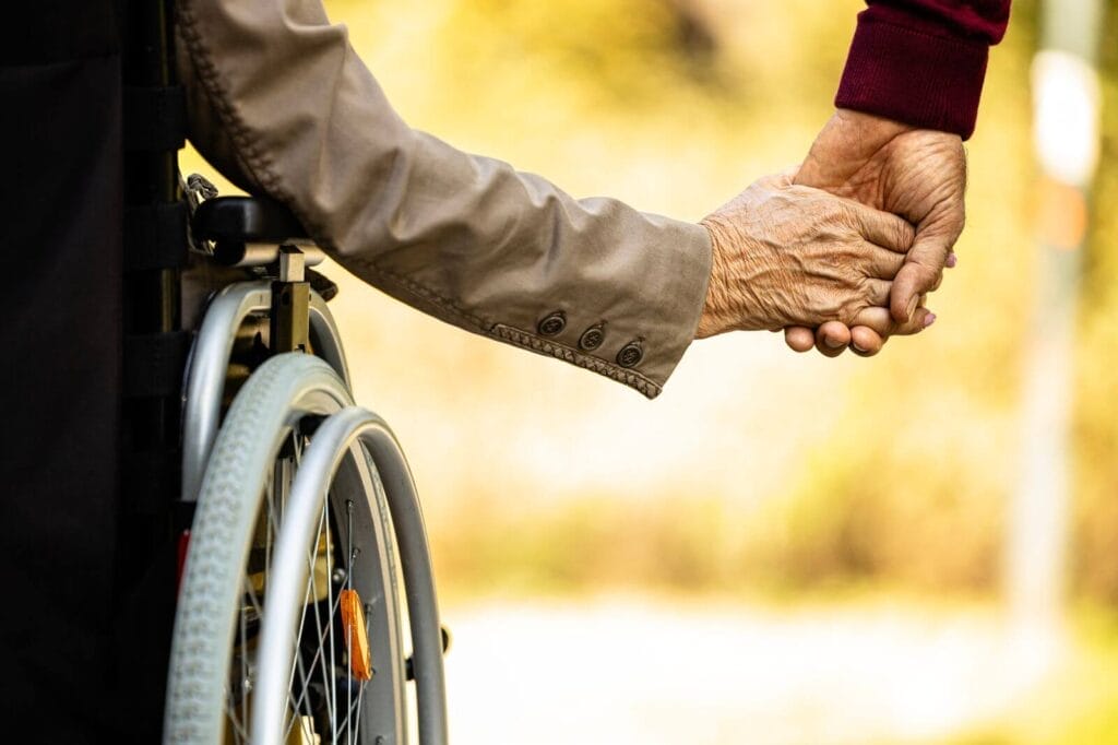 A close-up of two older adults' hand holding each other. One person is in a wheelchair.