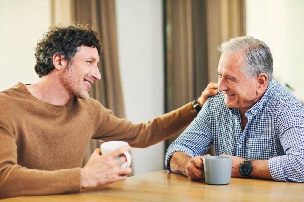 A man and his older adult father sit at a table drinking coffee and smiling at each other. The younger man has his hand on his father's shoulder.