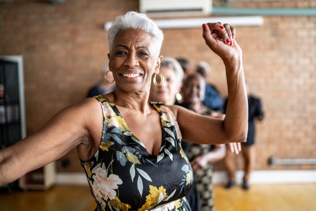 A smiling older adult woman is dancing while looking at the camera.