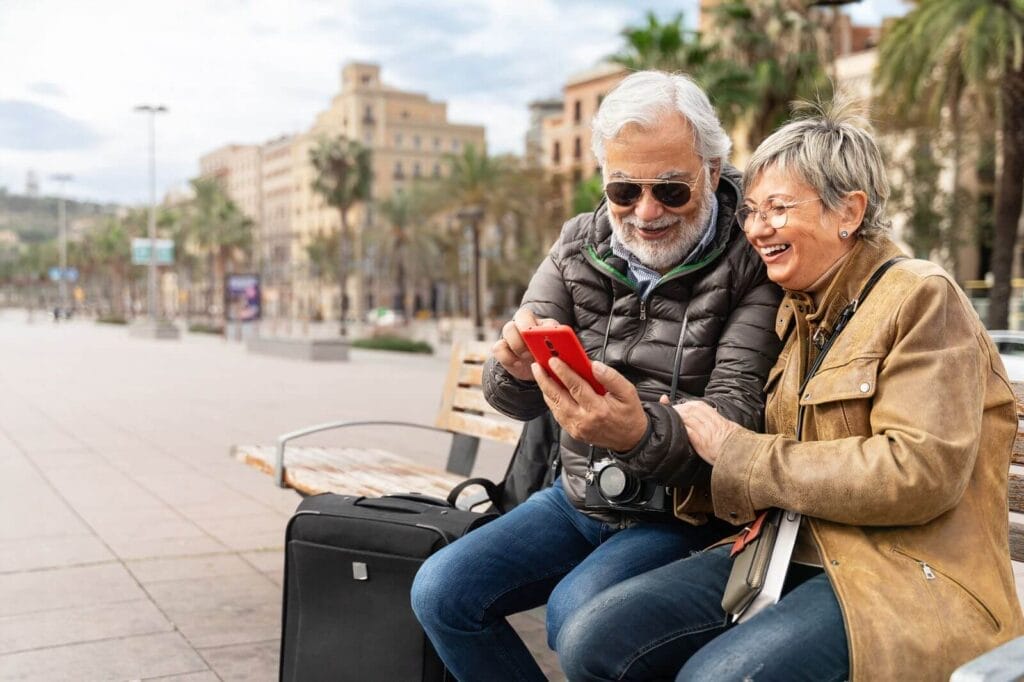 An older adult man and woman sit on a bench outside, looking at a cell phone. There is a suitcase next to the man.
