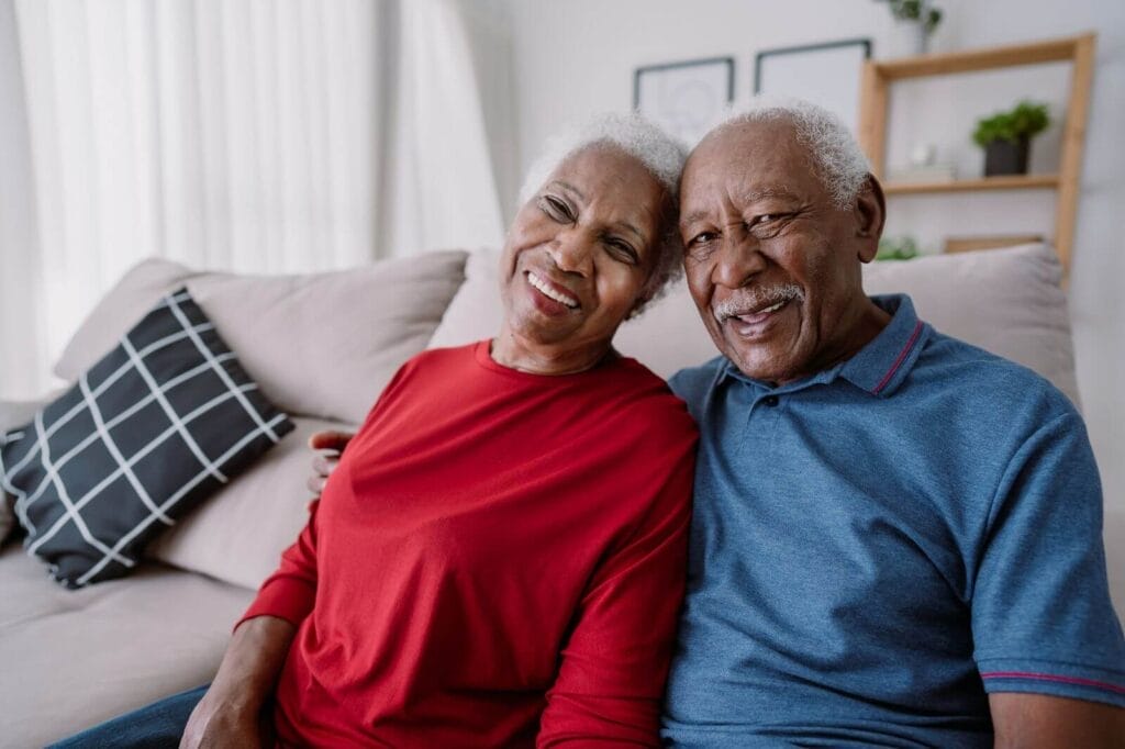 An older adult man and woman sit on a couch, smiling at the camera.