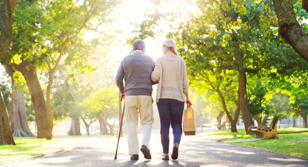 Shown from behind, a woman holds the elbow of an older adult man who is walking using a cane. They walk along a tree-lined path.