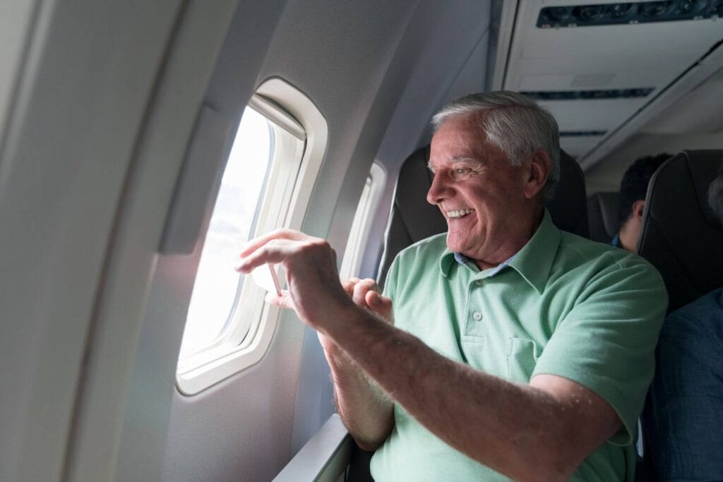 An older adult man looks out an airplane window, taking a picture with his cell phone.