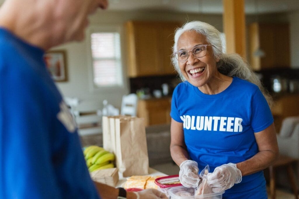 An older adult woman wearing a T-shirt that reads "Volunteer" is making sandwiches while smiling at a man across from her.