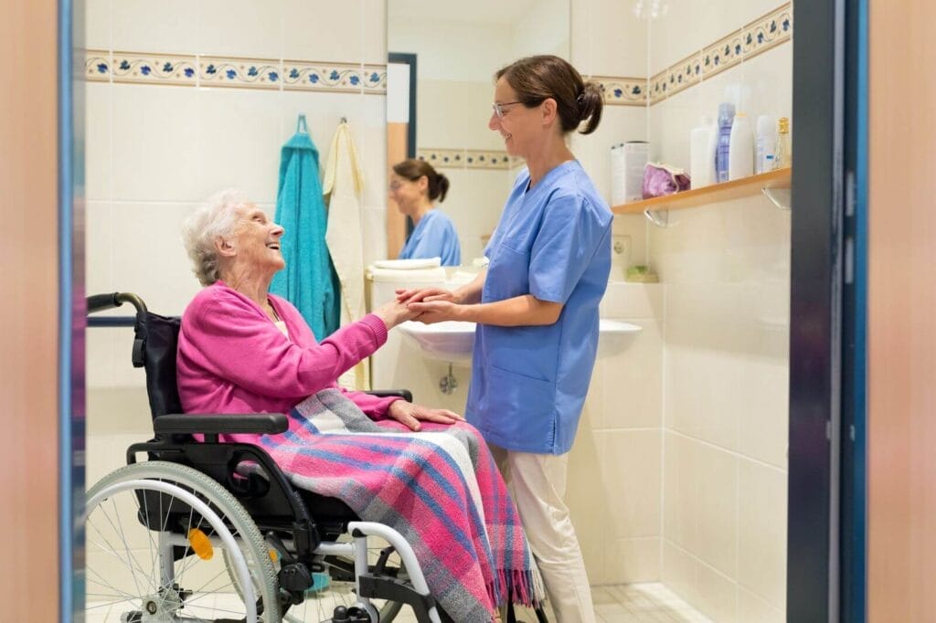 A female home care provider holds the hand of an older adult woman sitting in a wheelchair. They are in the bathroom in front of the sink.