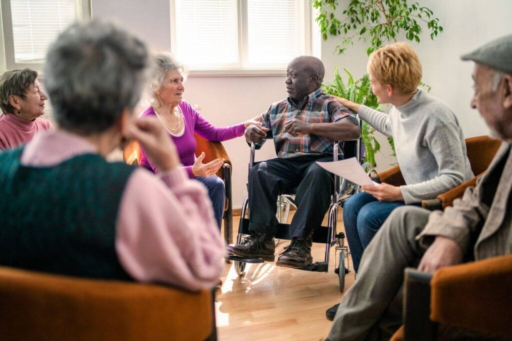 A group of six older adults sit in a circle. Two woman have their hands on the shoulders of a man. They are all talking.