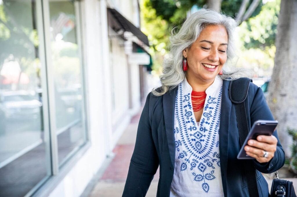 An older adult woman is walking outside, looking down at her cell phone and smiling.