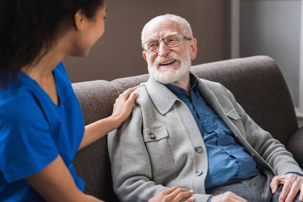 A female caregiver sits on a couch next to an older adult man with her hand on his shoulder. He smiles at her.