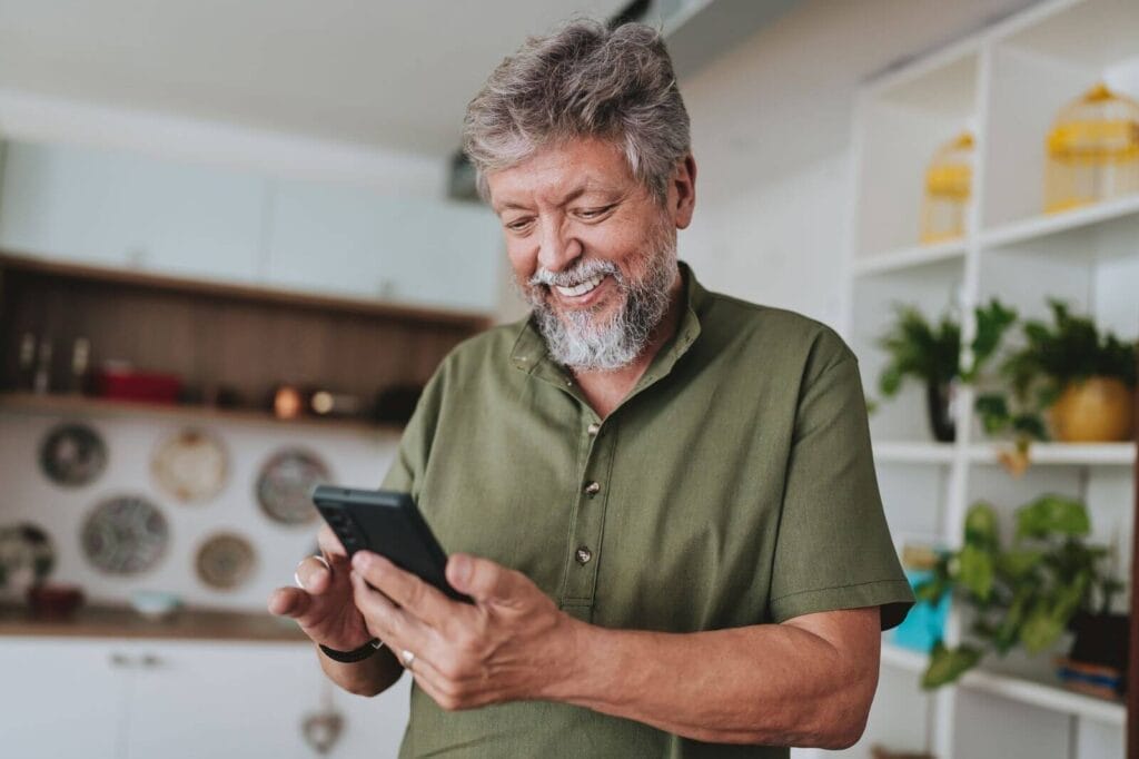 An older adult man is standing in his kitchen, looking down at his cell phone and smiling.