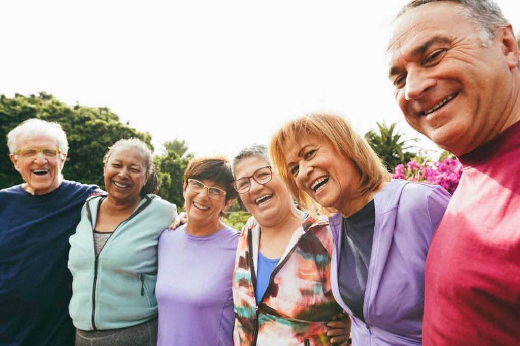 A group of six older adult men and women are standing outside with their arms around one another, looking at the camera and smiling.