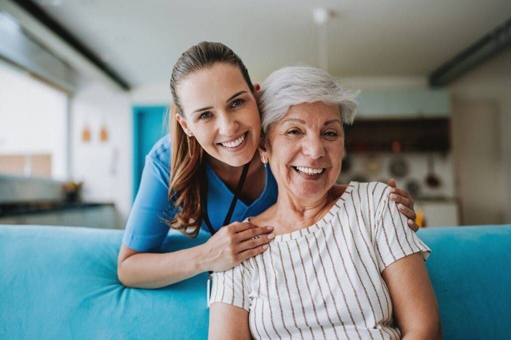 An older adult woman is sitting on a couch. A woman caregiver is bending over behind her to hold her shoulders in her hands. They both smile at the camera.