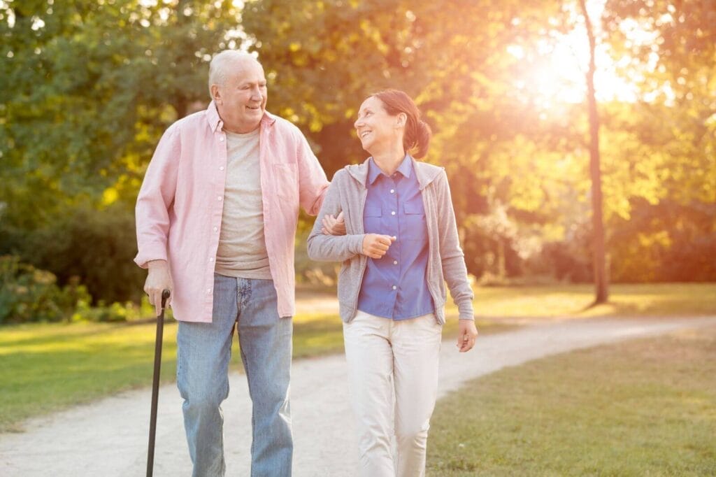 An older adult man using a cane is walking on a path outdoors, arm in arm with an older adult woman.