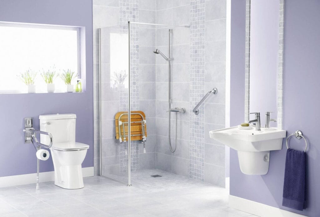 The image shows an empty bathroom with a sink, a walk-in shower with grab bars, a handheld showerhead, and a fold-down shower seat installed in the wall. There is also a toilet with a grab bar on one side.