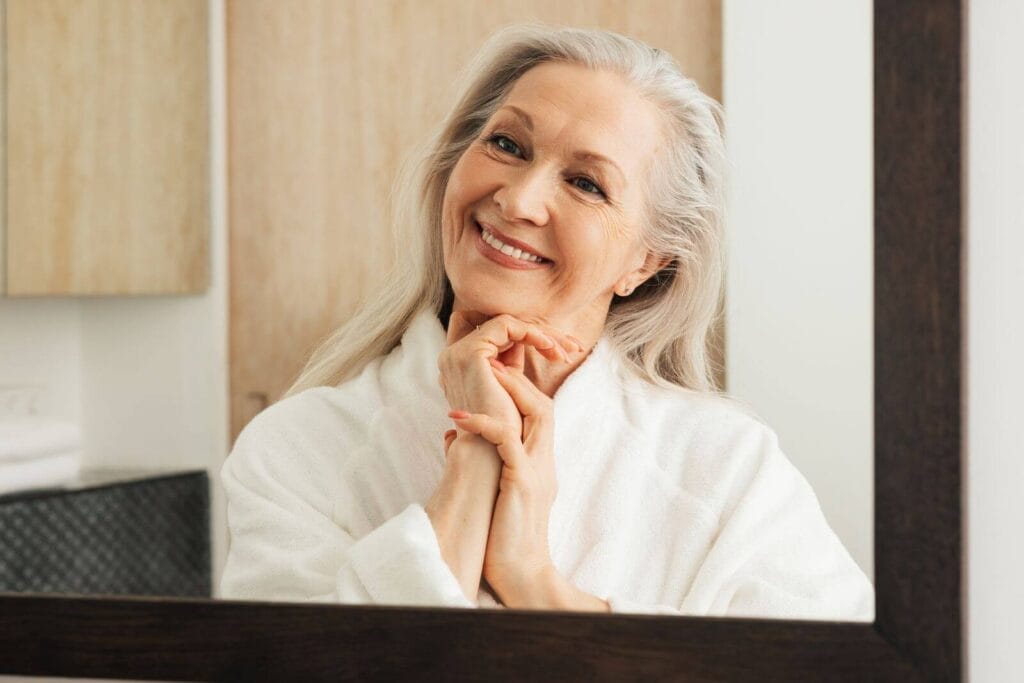 An older adult woman wearing a bathrobe smiles at herself in the bathroom mirror.