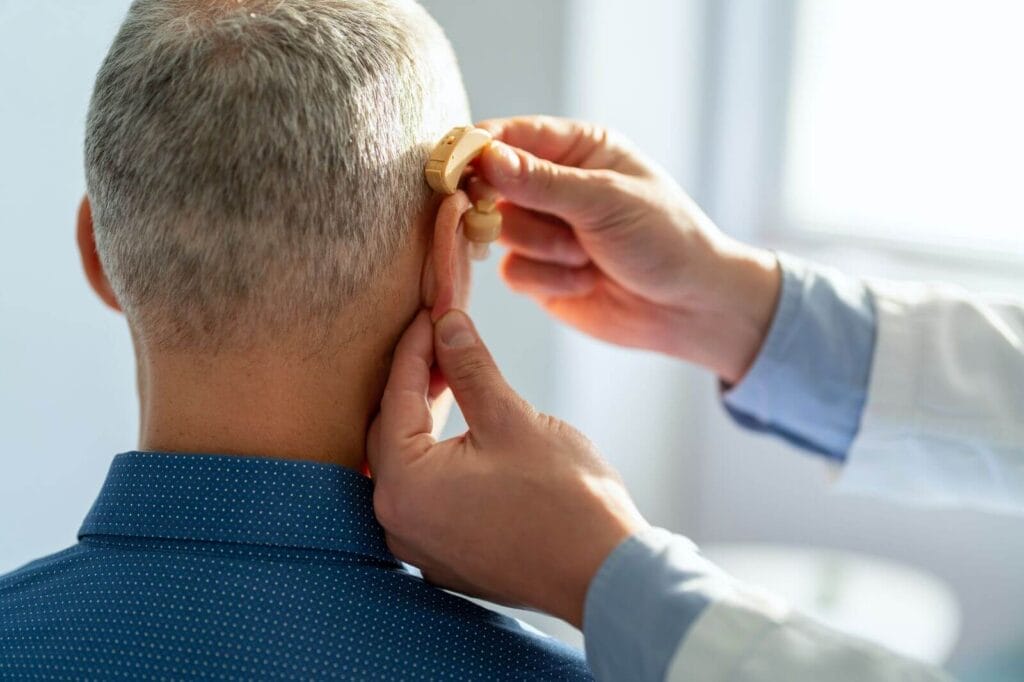A doctor places a hearing aid on the right ear of an older adult man.