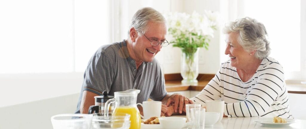 An older adult man and woman sit at a table with plates and mugs around them. They're holding hands and laughing.