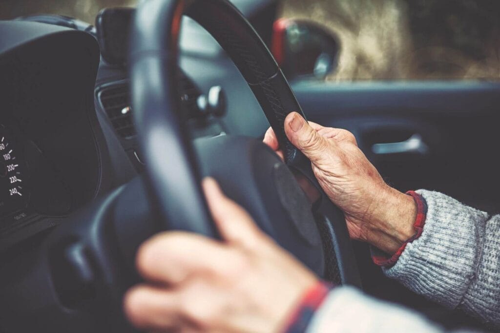 An older adult's hands hold the steering wheel of a car.