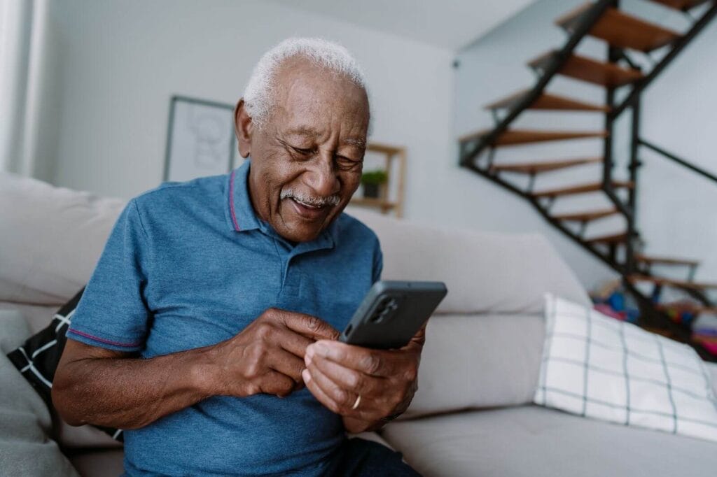 An older adult man sits on a couch, looking at his cell phone.