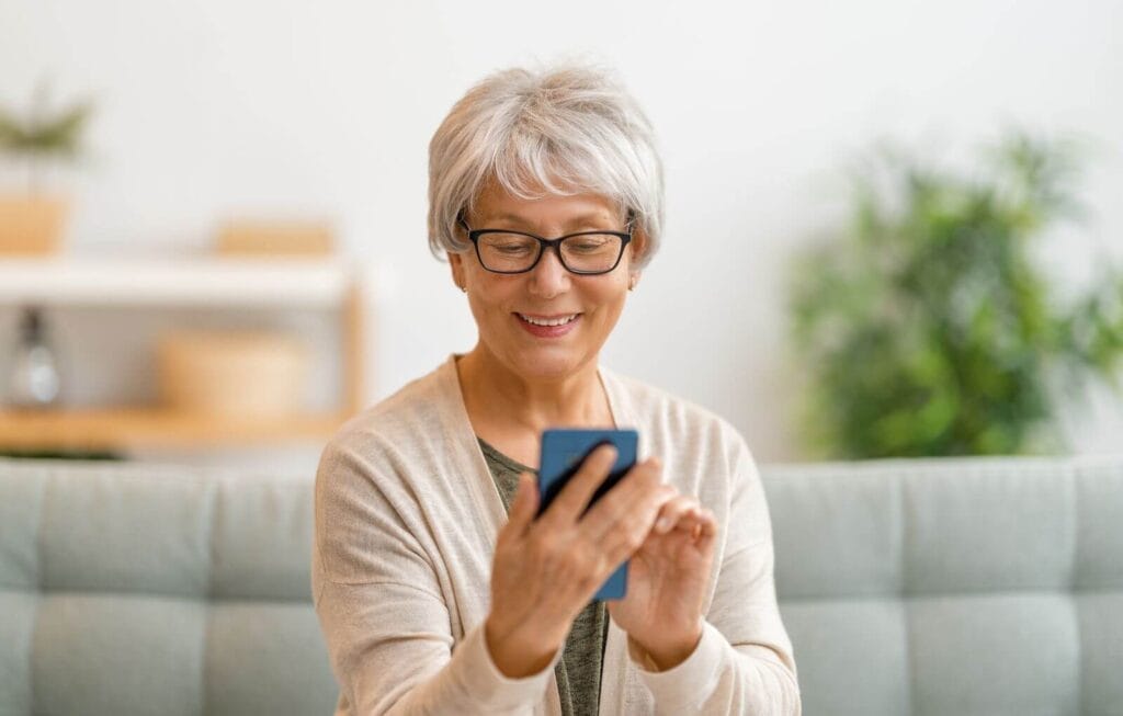 An older adult woman is sitting on a couch, looking down at her cell phone and smiling.