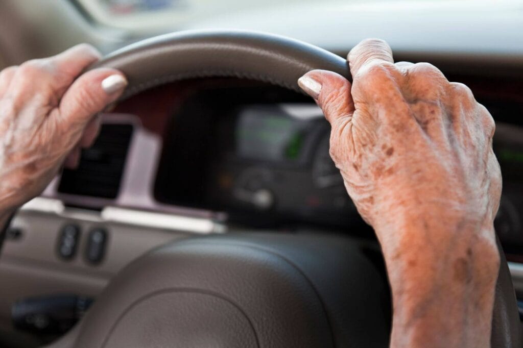 An older adult woman's hands hold a car's steering wheel.