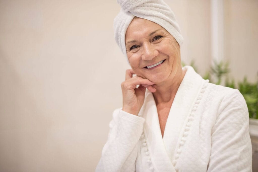 An older adult woman wearing a bathrobe and a towel wrapped around her hair smile at the camera while holding her hand to her face.