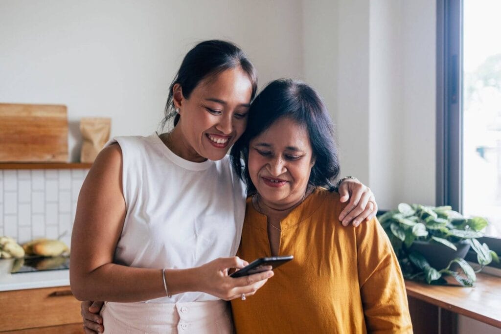 A woman stands next to an older adult woman, showing her her cell phone. They smile and have their arms around each other.