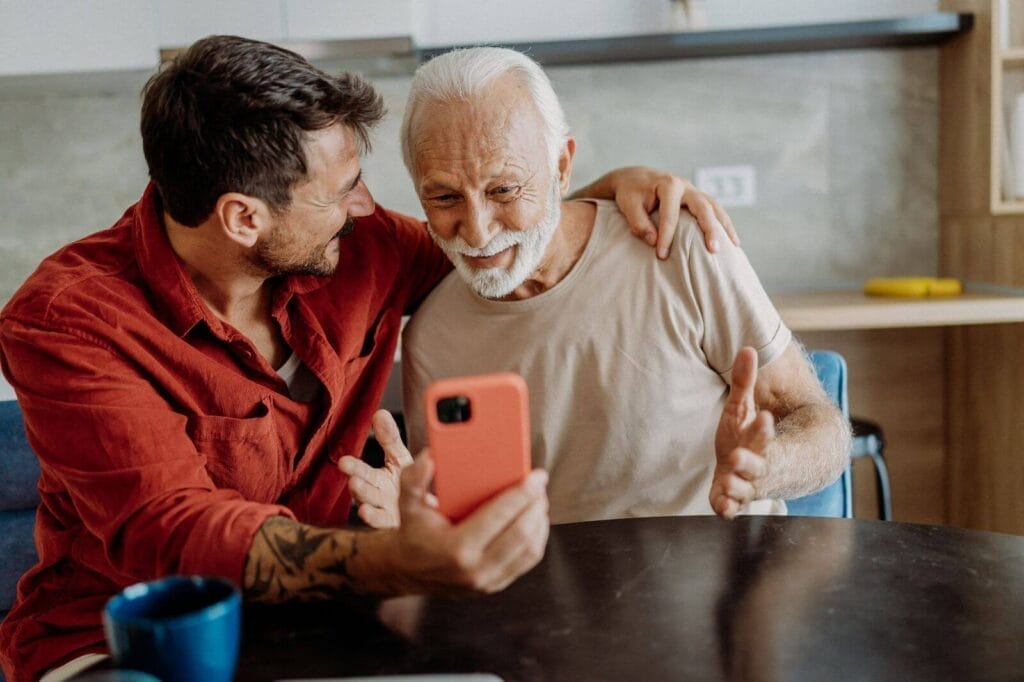 An older adult man and a younger man sit at a table. The younger man is holding a cell phone and has his arm around the older man's shoulders. They are smiling.