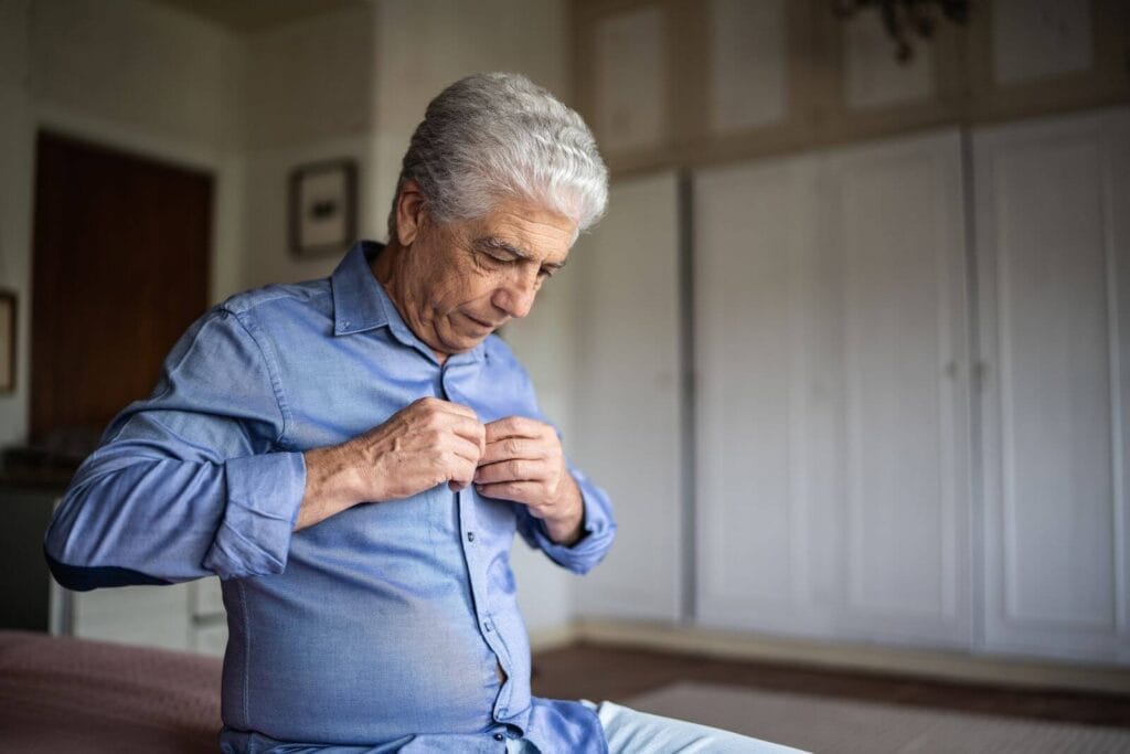 An older adult man sits down, buttoning his shirt.