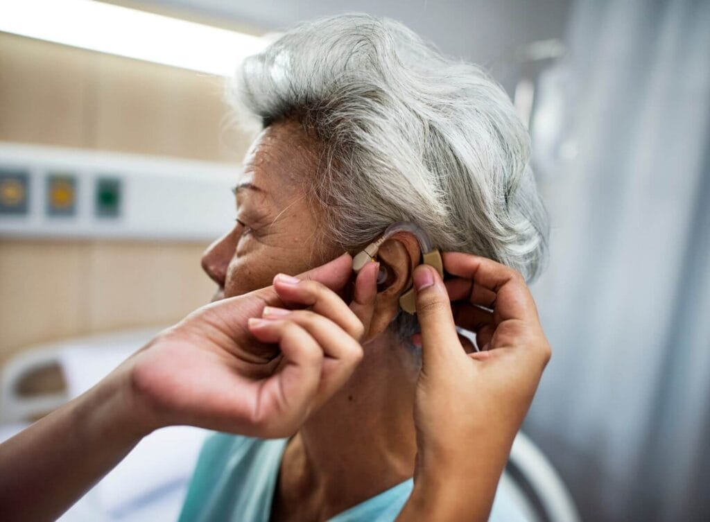 A person's hands are shown placing a hearing aid on an older adult woman's ear.