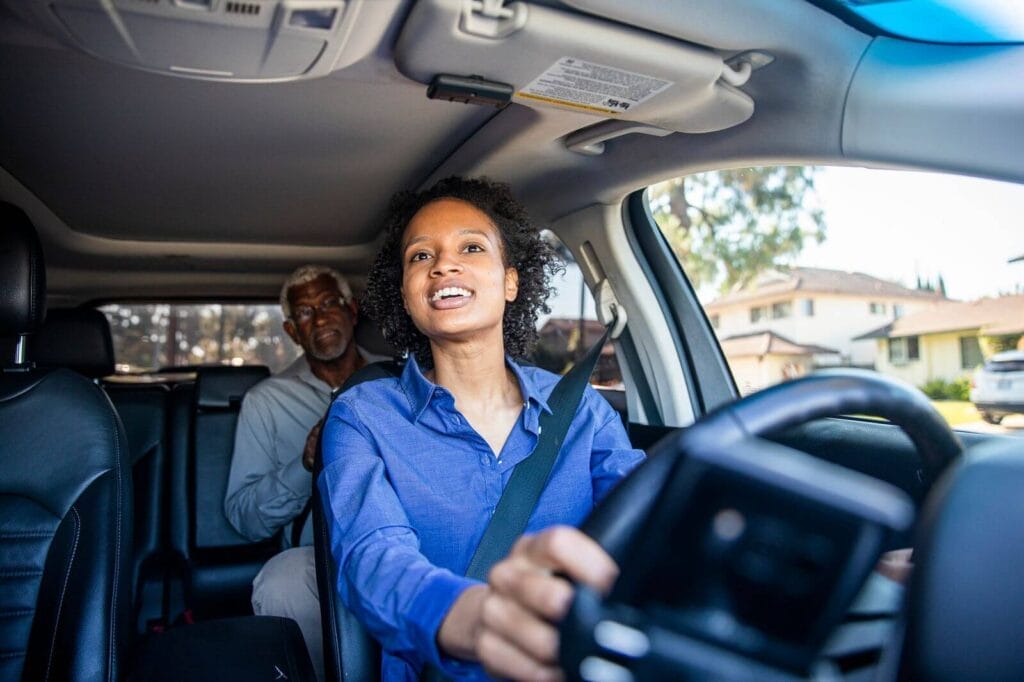 A woman looks through her rearview mirror at an older adult man who is seated in the back seat of her car.