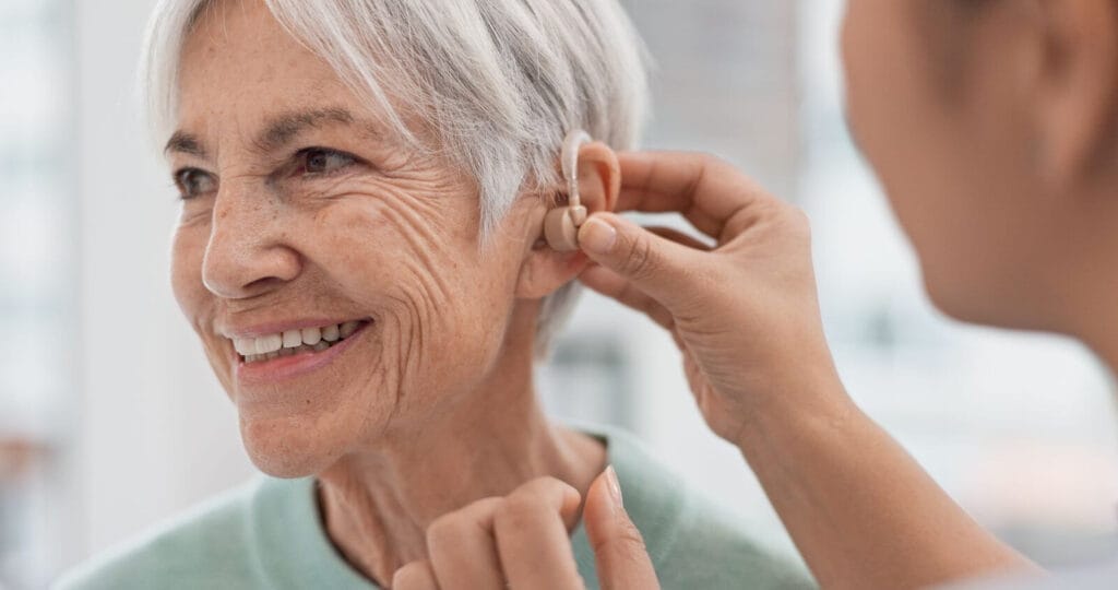 An older adult woman smiles as a person whose face is off-camera places a hearing aid in her ear.