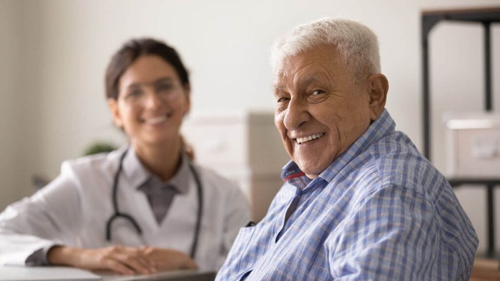 An older adult man smiles at the camera with a younger woman doctor in the background.