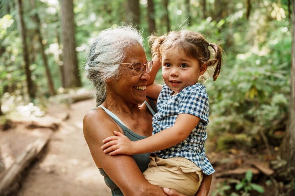 An older adult woman holds her young granddaughter in her arms. They are outside on a trail in the woods.