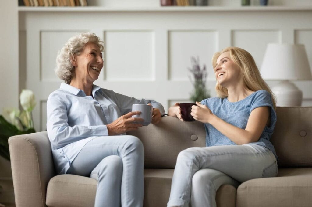 A woman and an older adult woman sit on a couch, drinking coffee and laughing at each other.