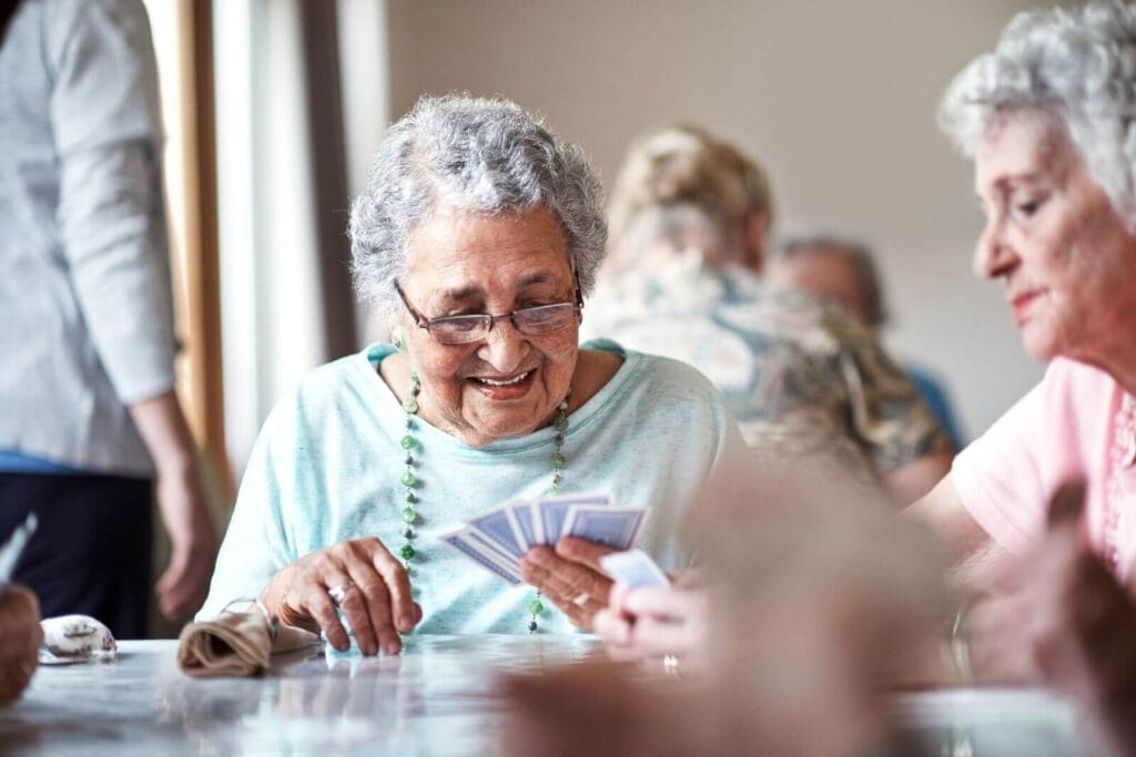 Two older adult women sit at a table playing cards. There are other older adults in the background at another table.