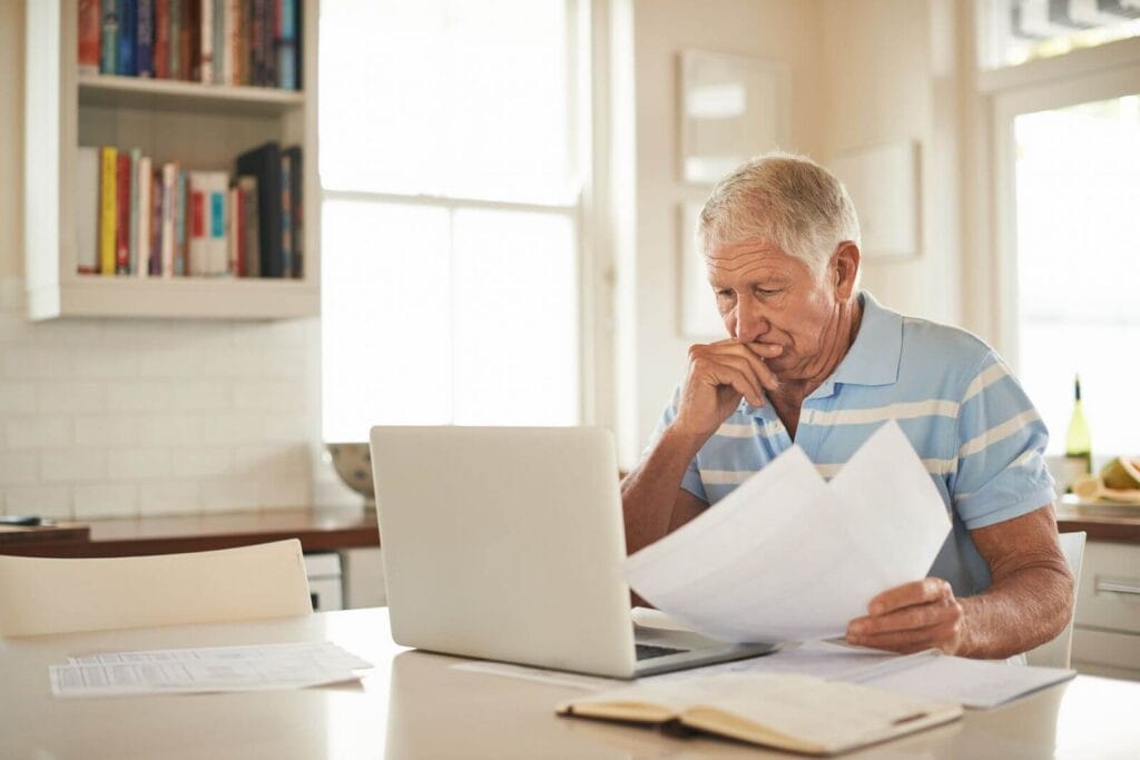 An older adult man sits at a table looking over paperwork. His laptop is open in front of him.