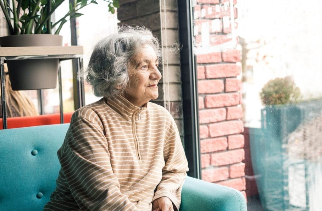 An older adult woman sits on a couch, looking out a window.