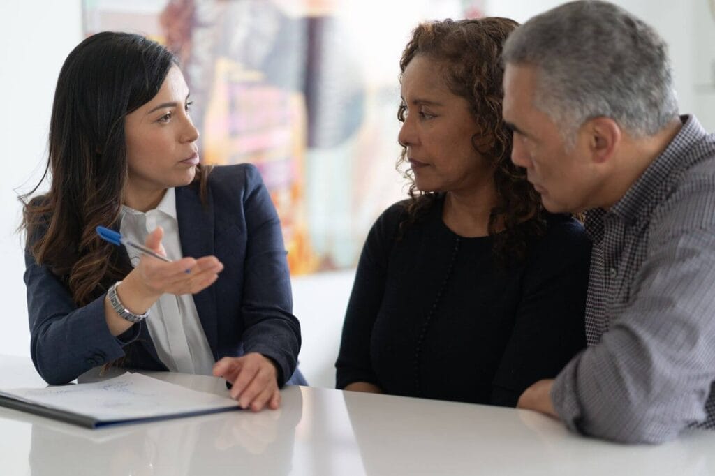 An older adult man and woman sit at a table with a younger woman discussing business. There is paperwork on the table in front of the younger woman.