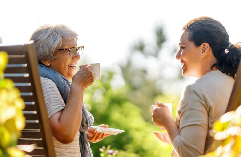 A woman and an older adult woman sit on chairs outside, drinking tea and smiling at each other.