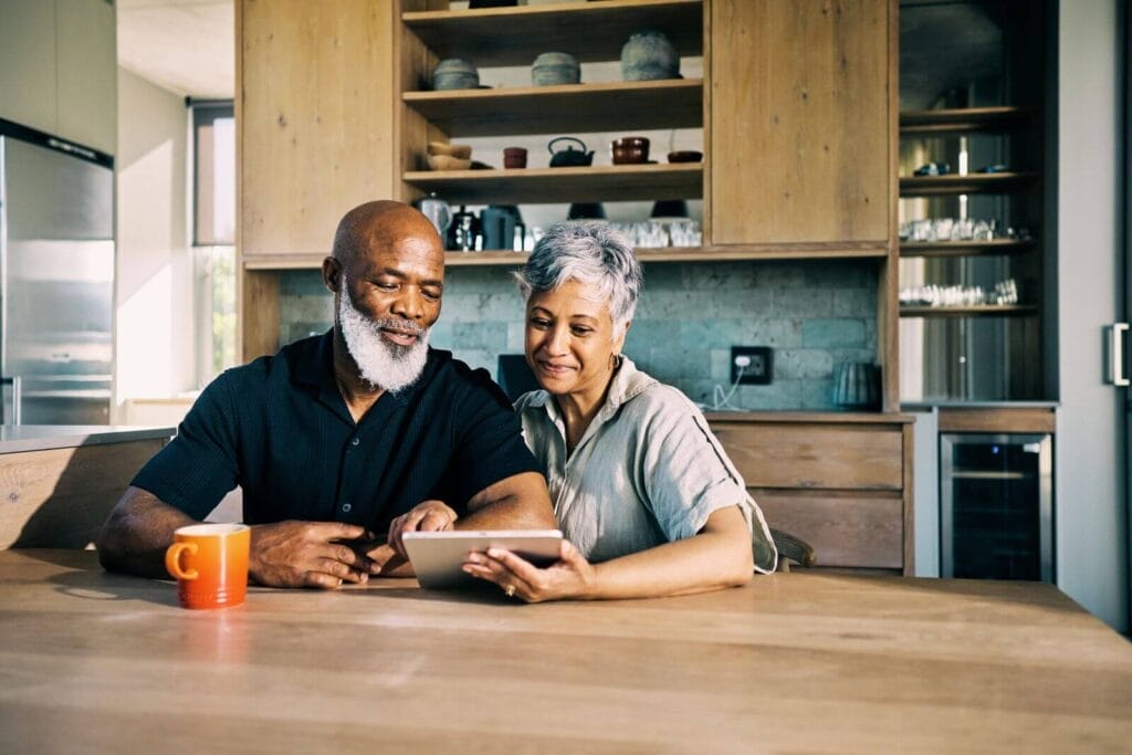 An older adult man and woman sit at their kitchen table looking at a tablet. There is a coffee mug in front of the man.