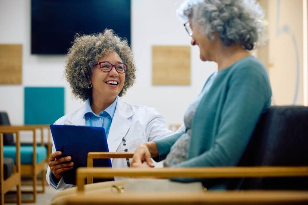 An older adult woman sits in a chair in a doctor's office waiting room. A woman doctor crouches next to her, holding a clipboard. They smile at each other.