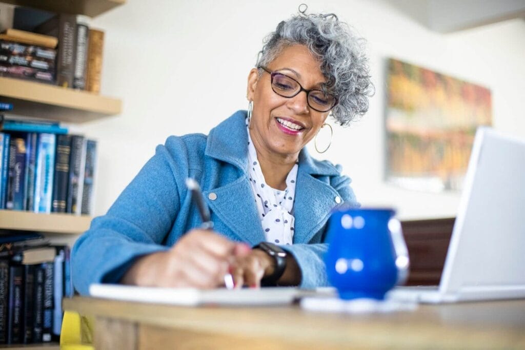 An older adult woman sits at a table in front of a laptop. She is taking notes in a notebook.