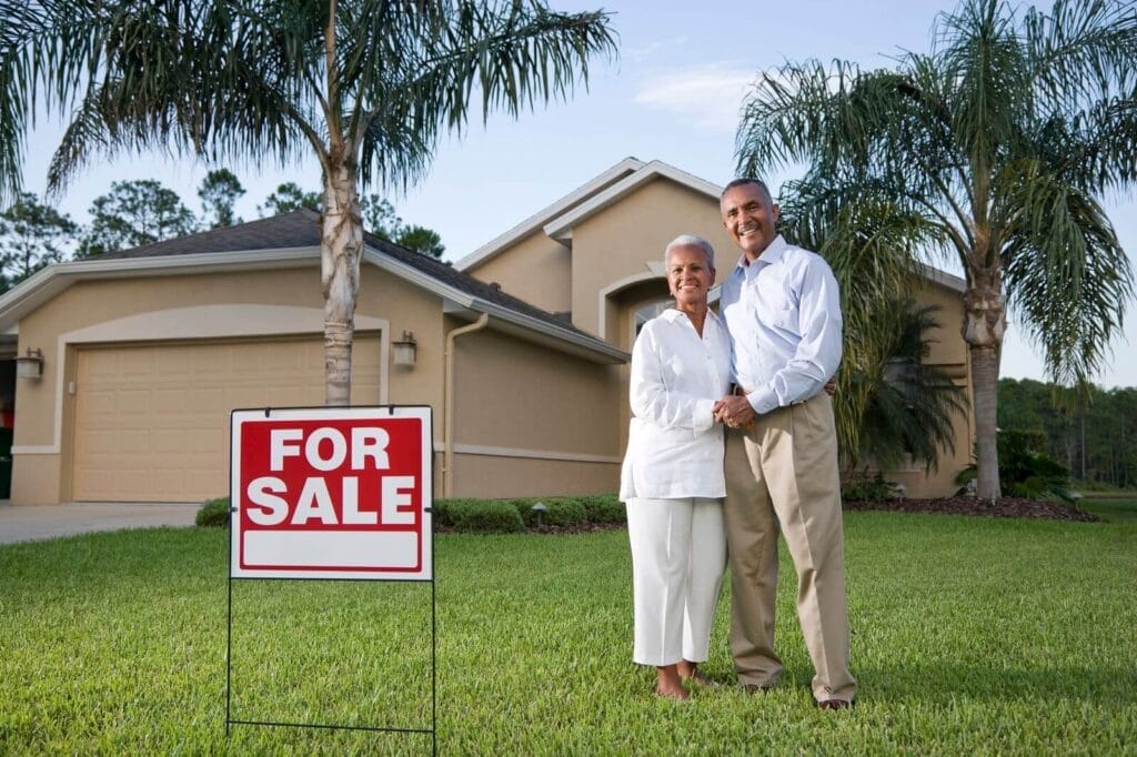 An older adult man and woman stand in front of a house with their arms around each other. They are next to a for sale sign.