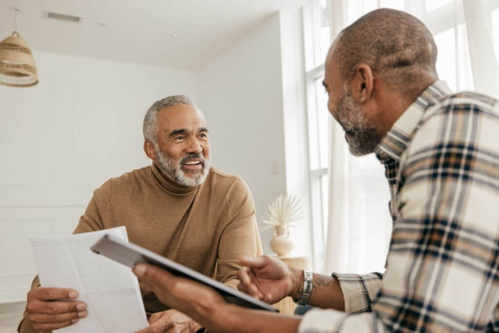 An older adult man and his adult son sit in chairs, holding paperwork and smiling at each other.