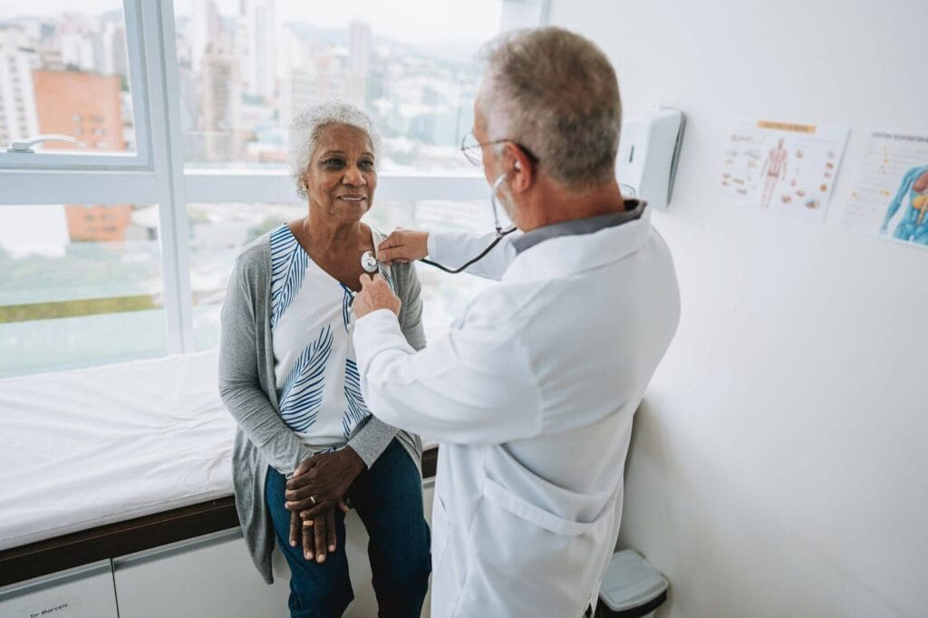 An older adult woman sits on an examination table at a doctor's office while a male doctor holds a stethoscope to her chest.