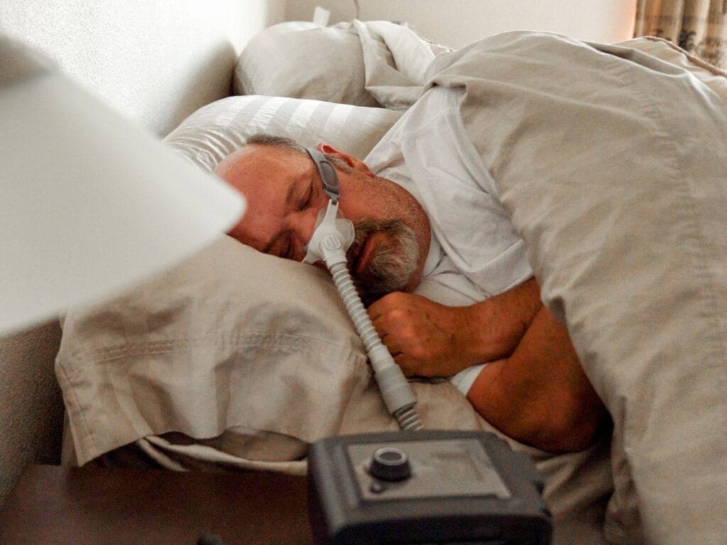 An older adult man sleeps in bed. He is using a CPAP machine with a nasal mask.