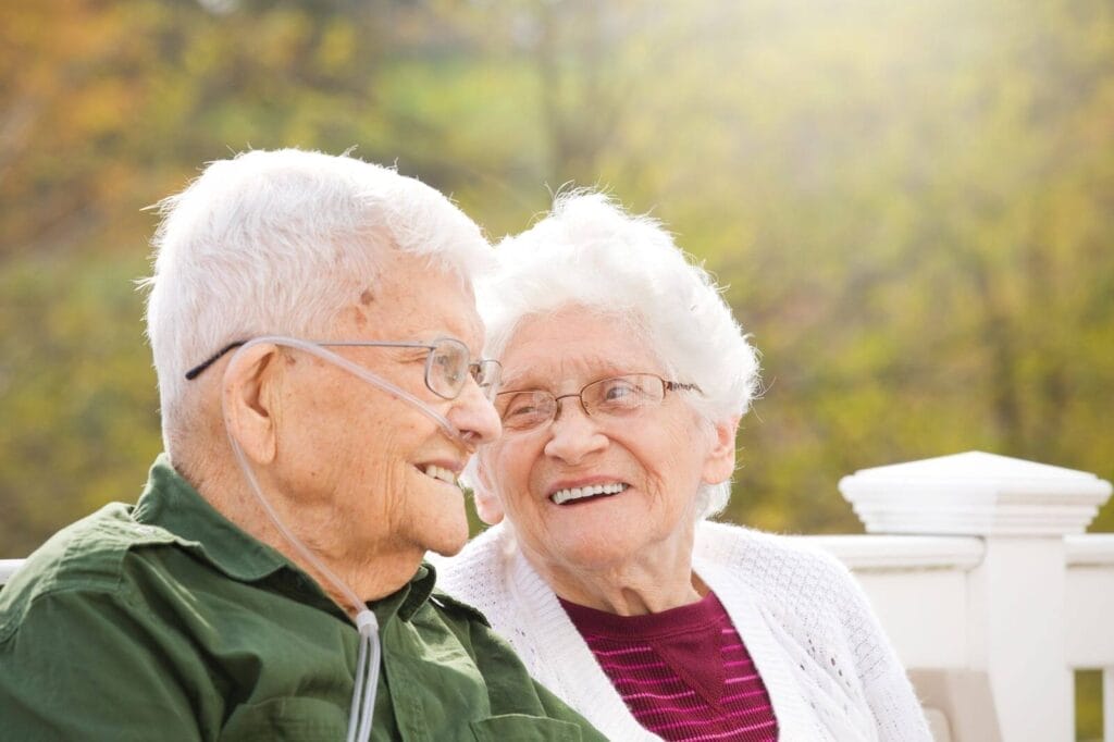 An older adult man and woman sit outside. The man is using a nasal cannula.